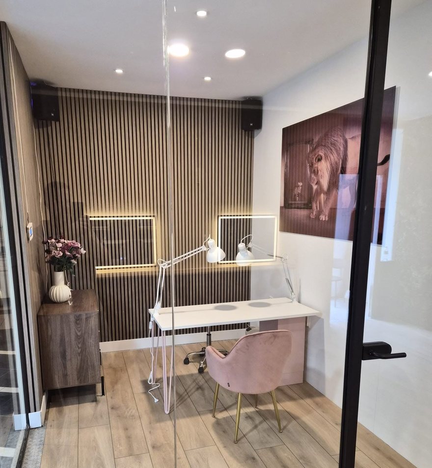 De werkplek van Glam & Glow Studio: een afgesloten ruimte met glazen wand, houten lattenwand, witte manicuretafel en roze fauteuil